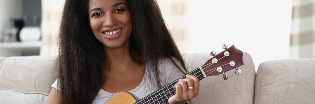Happy afro american woman playing on ukulele in living room at home