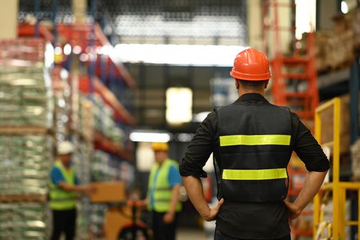Back view of male manager wearing hardhat and reflective jacket checking inventory production stock control in warehouse