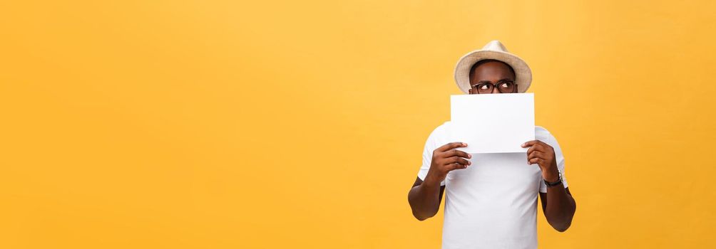 Young happy African-american hiding behind a blank paper, isolated on yellow background
