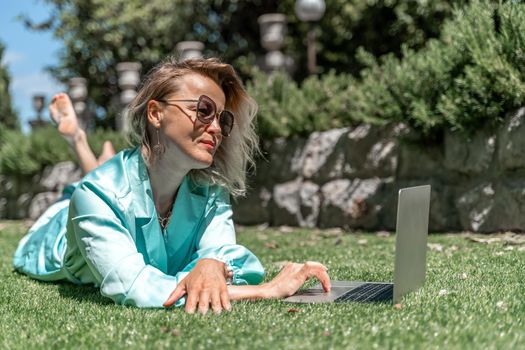 a young beautiful woman with blond curly hair in glasses and a blue dress sits on the grass in nature and uses a laptop,