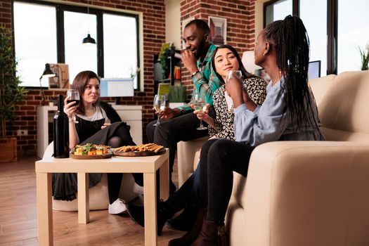Culturally diverse social gathering of people drinking wine, talking, chatting, enjoying themselves, eating snacks at house made party. Diverse friends enjoying partnership.