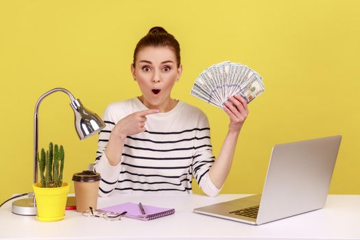 Shocked woman pointing at hundred dollar bills, holding sitting at workplace with laptop high salary
