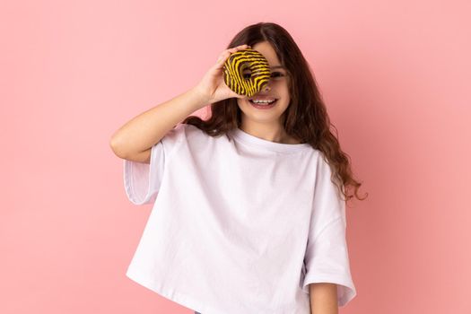 Funny adorable little girl standing having fun, holding and covering eye with donut and smiling.
