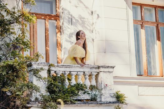 a beautiful smiling and kind woman in a gorgeous yellow dress stands on the balcony of an old vintage house