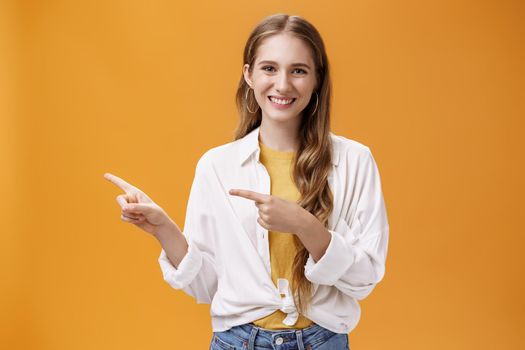 Waist-up shot of friendly pleasant and stylish young teenage female in blouse over t-shirt with cute wavy fair hair pointing left and smiling broadly at camera posing happy against orange background