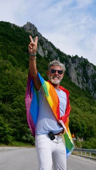 Portrait of a gray-haired senior elderly Caucasian man bisexuality with a beard and sunglasses with a rainbow LGBTQIA peace flag in mountains. Celebrates Pride Month, Rainbow Flag Day, gay parade