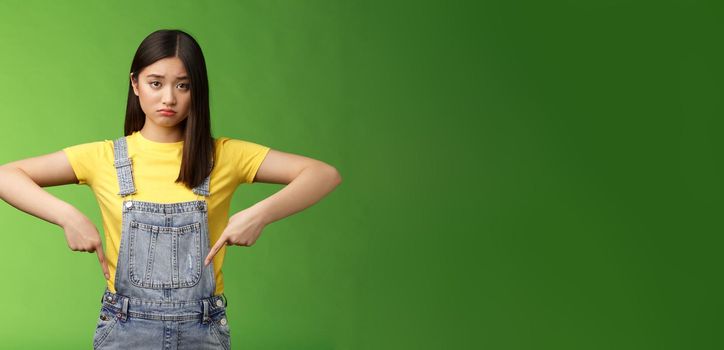 Sad asian girl in dispair feel upset, frowning sulking pessimistic, pointing down look camera pulling sorrow uneasy face, express sadness and frustration, stand green background