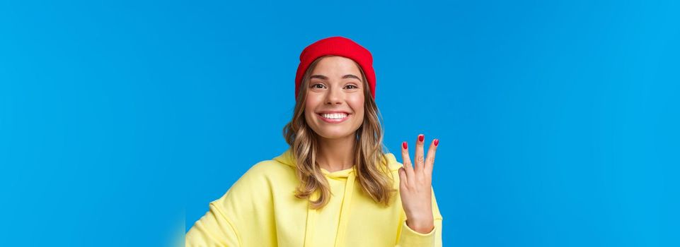 Good-looking caucasian female in red beanie and yellow hoodie, showing number three and smiling, make order or reservetion, standing proud and delighted, blue background