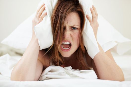 Morning already. Portrait of an angry young woman shouting while lying in bed with a pillow over her head.
