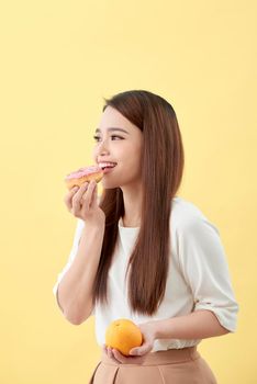 Dieting concept. Beautiful Young Asian Woman holding orange and donut over yellow background. Choosing between junk food with healthy food.