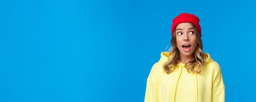 Excited good-looking caucasian female student in red beanie and yellow hoodie, look upper right corner and open mouth fascinated, hear interesting gossip, standing blue background