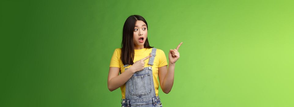 Shocked gasping alarmed pretty asian girl drop jaw impressed, turn pointing left hear loud bang, stand astonished speechless, gazing curious event, pose green background