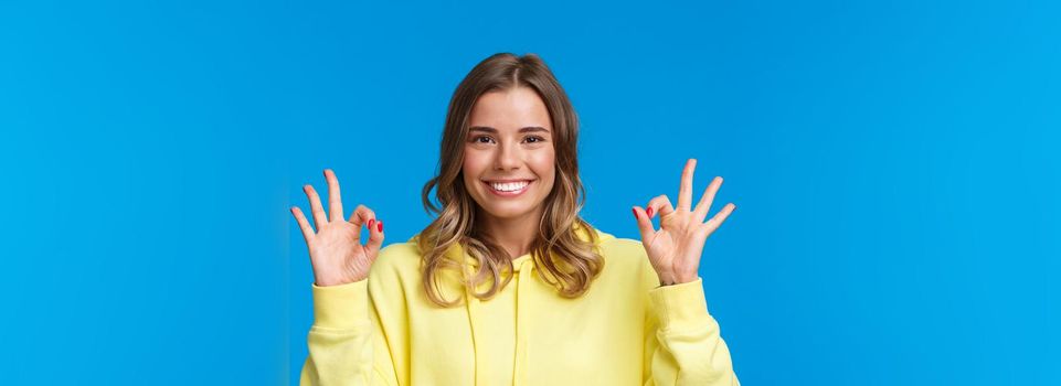 Close-up portrait happy confident young blond girl assure all okay, guarantee plan went well, smiling and show ok gesture in approval, confirmation or like, stand blue background