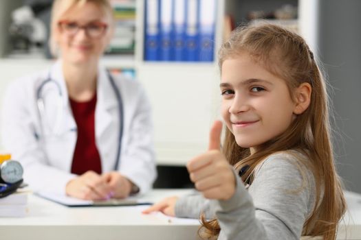 Beautiful little girl shows thumbs up. and doctor sits in background