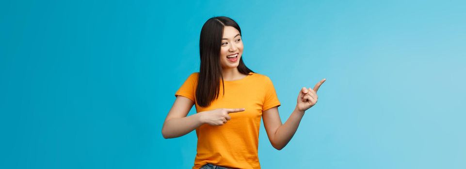 Friendly enthusiastic asian girl curiously looking sideways, pointing left interesting object, grinning joyfully discuss new opened store, stand blue background carefree lively talking