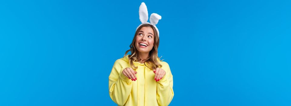 Holidays, traditions and celebration concept. Dreamy and cute young woman smiling lovely with rabbit ears, look upbeat upper right corner grinning make paws gesture, blue background