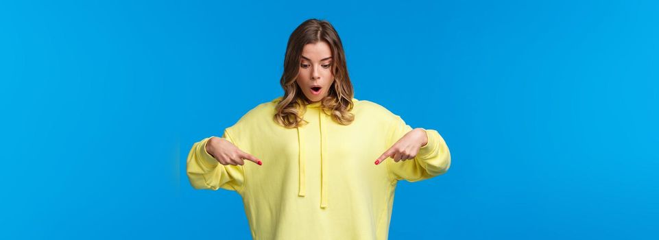 Girl found something stunning. Amused and speechless surprised blond alluring woman in yellow hoodie, drop jaw stare and pointing fingers down at cool product, blue background