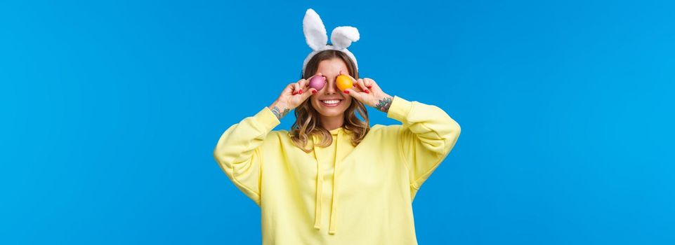 Holidays, traditions and celebration concept. Happy carefree smiling caucasian woman celebrating Easter, holding two painted eggs on eyes and grinning, wear cute rabbit ears, blue background