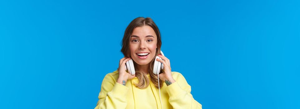 Close-up portrait of excited happy smiling blond girl in yellow hoodie, take-off headphones and grinning as meet friend on street stop by for conversation, standing joyful blue background