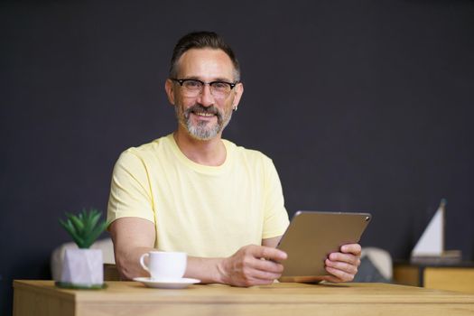 Working from home middle aged man use digital tablet sitting at home office desk wearing casual with black wall on background. Handsome freelancer mature man hold tablet pc working from home