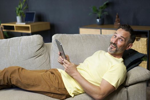 Laying on the couch middle aged freelancer man use digital tablet to read social media or working offline from home wearing casual. Relaxed freelancer man laying on sofa making business uses gadget