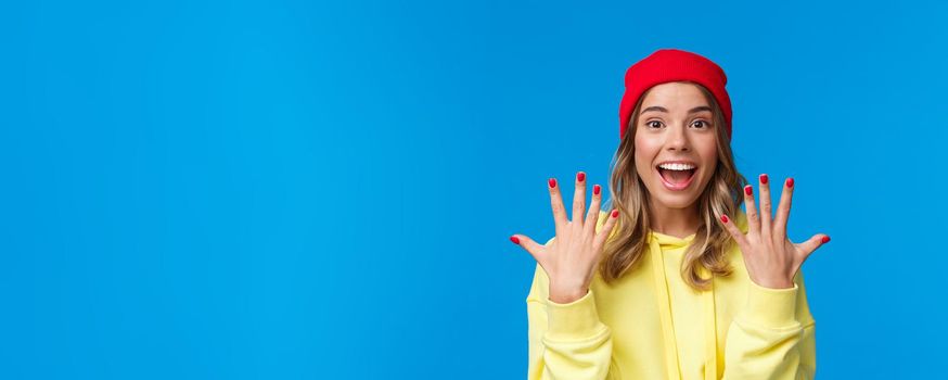 Portrait of amused good-looking blonde hipster girl smiling joyfully, explain something showing number ten, tenth with hands and look camera upbeat, order dozen, stand blue background