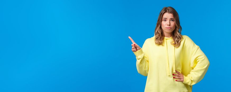 Upset and insecure cute european blond young woman in yellow hoodie, having doubts, being skeptical about topic, pointing finger upper left corner and look unsure camera, blue background