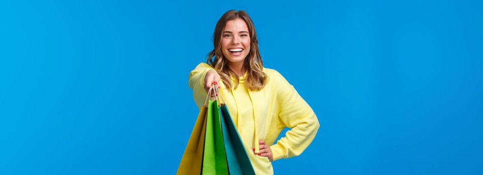 Cheerful blond girl giving you shopping bags and smiling happy, buying presents for family holiday, buying lots of staff in store and asking carry her packages, stand blue background