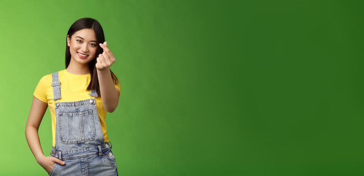 Lovely tender carefree asian girlfriend show korean love sign, make finger heart smiling cute, showing passion and affection, stand green background in overalls and yellow t-shirt, express sympathy