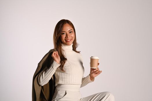Portrait of modern young woman holding paper cup, sitting on white background. Studio photo, Autumn and Winter concept