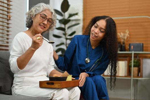 Smiling female caregiver serving dinner for elderly woman. Home health care service concept.