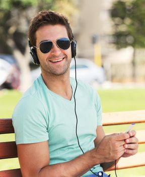 Beats on a bench - inner city living. A smiling young man wearing sunglasses sitting on a bench in a park and listening to music through his headphones.