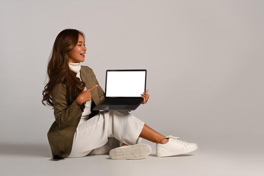 Stylish young woman pointing on laptop screen, sitting on white background. Blank screen for your advertise text