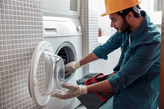 Focused repairman in worker suit is fixing washing machine in bathroom
