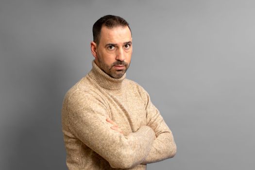 Bearded hispanic man dressed in brown turtleneck sweater standing defiantly with crossed arms looking serious at the camera isolated over gray background.