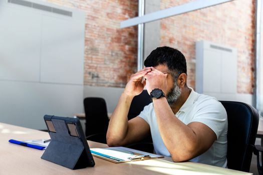 Mid adult Indian man worried and overwhelmed with work, forehead resting on hands at the office. Copy space.