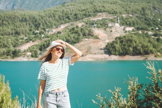 Smiling woman in hat and sunglasses with wild hair standing near mountains lake on background. Positive young woman traveling on blue lake outdoors travel adventure vacation