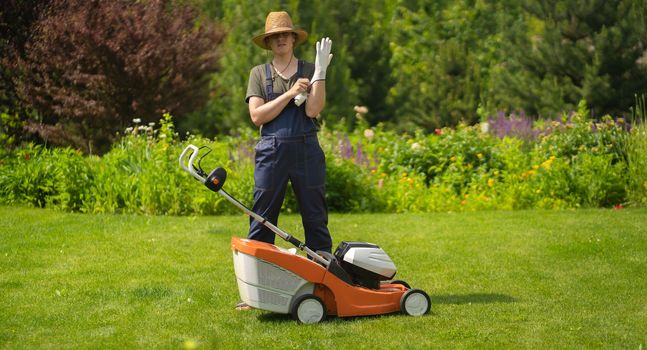 A young man in a straw hat is putting on gloves to mowe a lawn with a lawn mower in his beautiful green floral summer garden.