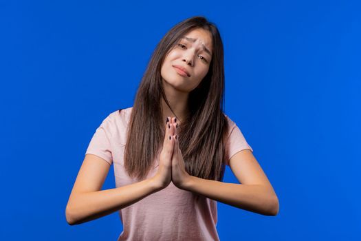 Young woman praying, begs to apologize her, she is guilty. Blue background. Hopeful prayer girl asking someone satisfy her desires, help with.