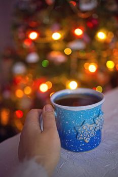 A woman's hands in a sweater hold a red cup of coffee by the window on the background of Christmas tree. Christmas lights. Hot winter flavored drink.