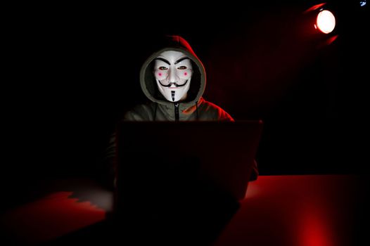 June 5, 2022 Novosibirsk, Russia: Anonymous in a hood is typing on a laptop in the dark in red light.