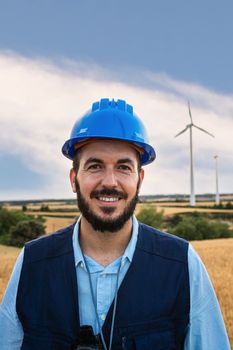 Vertical portrait of happy electric engineer standing at wind turbine farm looking at camera. Renewable energy.