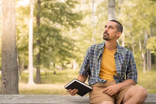 Young man sitting outdoors holding book in a park with trees background - Knowledge faith concept.