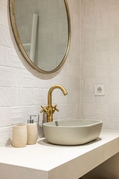 Oval stylish sink on white smooth marble countertop in modern bathroom. Copper-colored two-valve faucet blends harmoniously with round mirror hanging on wall and other objects in room.
