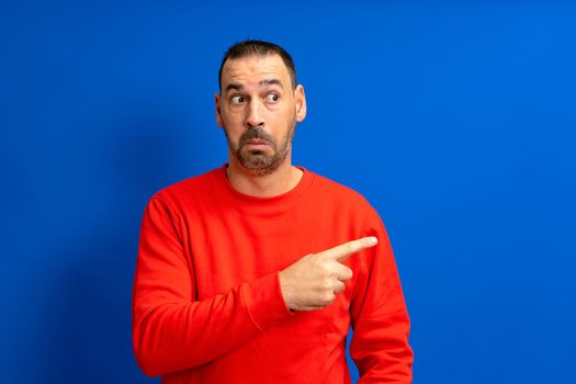 Bearded hispanic man dressed in red sweater pointing to the side with index finger while looking to the side skeptically, isolated on blue studio background.