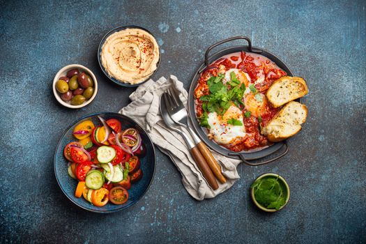 Middle Eastern breakfast or brunch with Shakshouka in pan, toasts, vegetables salad, hummus, olives