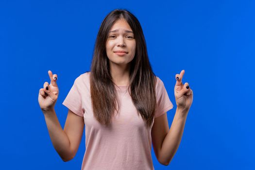 Pretty teen girl praying with crossed fingers on blue background. Woman begs someone satisfy her desires, help with, prays for her luck in exam