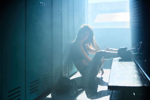 Young girl of an athletic build in the locker room of a sports club wears boxing gloves, a girl is dressed in sports black underwear, sun rays from window, sneakers