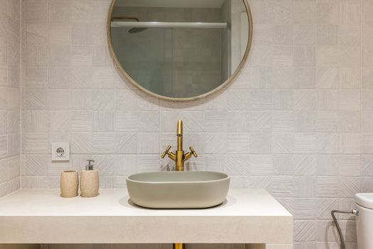 Oval stylish sink on white smooth marble countertop in modern bathroom. Copper-colored two-valve faucet blends harmoniously with round mirror hanging on tiled wall