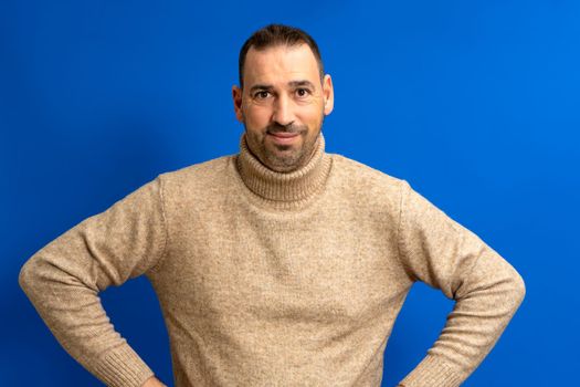 Cheerful bearded man wearing a turtleneck sweater posing isolated over blue background, studio portrait. People lifestyle concept. Mock up copy space. Standing with arms akimbo on his waist blinking.